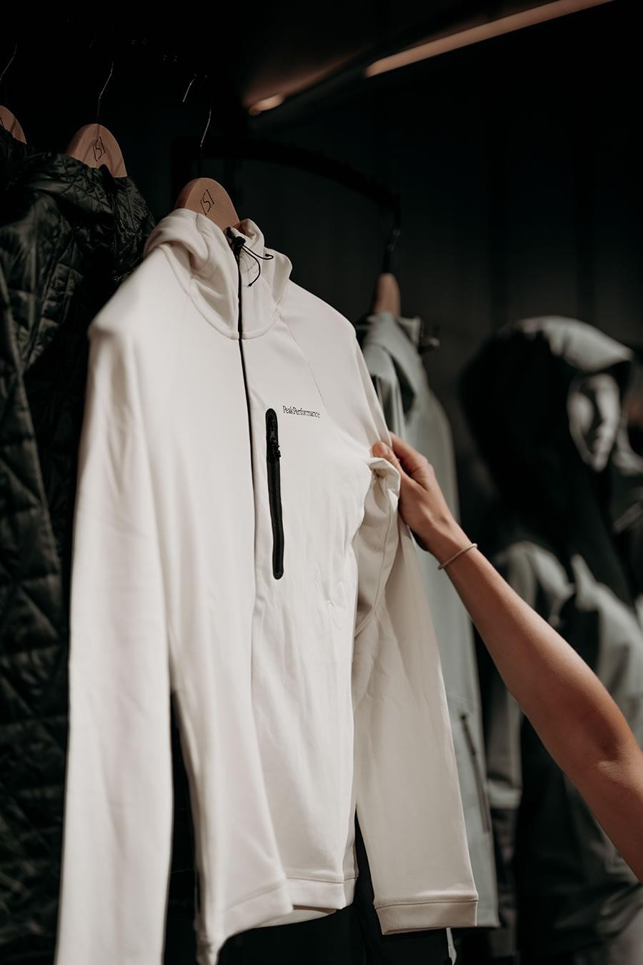 Die linke Hand hält einen weißen Pullover mit schwarzem Reißverschluss auf einem Bügel