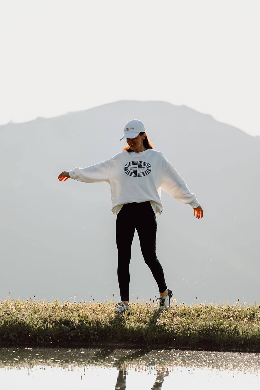 Person in weißem Pullover und Mütze steht auf Wiese, reflektiert im Wasser