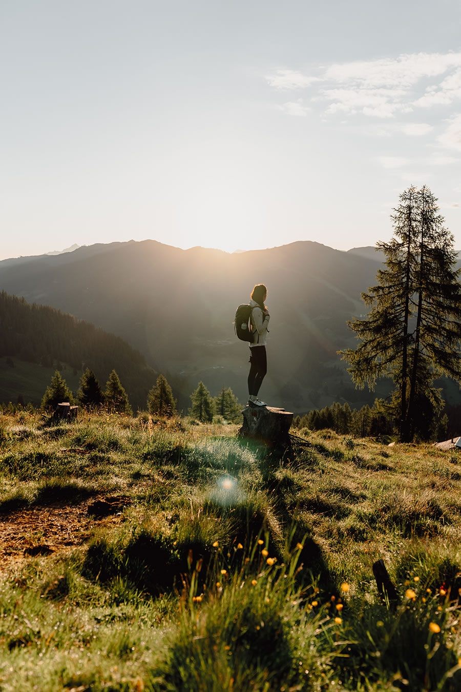 Eine Person mit Rucksack steht bei Sonnenuntergang auf einem Baumstumpf in einer Berglandschaft