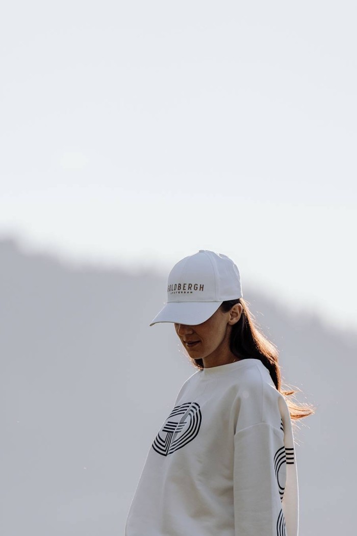 Frau mit weißem Cap und Pullover steht im Sonnenlicht vor unscharfem Hintergrund