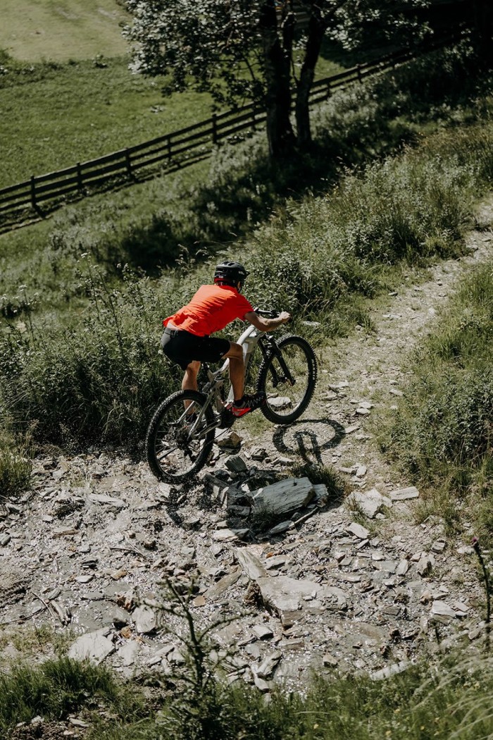 Person in rotem Shirt fährt mit Mountainbike auf einem schmalen, steinigen Pfad bergauf