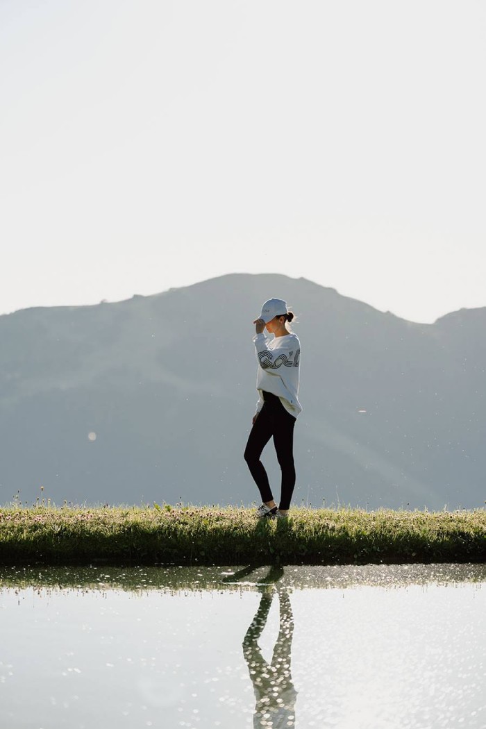 Person spaziert auf einer Wiese vor einem Berg, mit Reflexion im Wasser