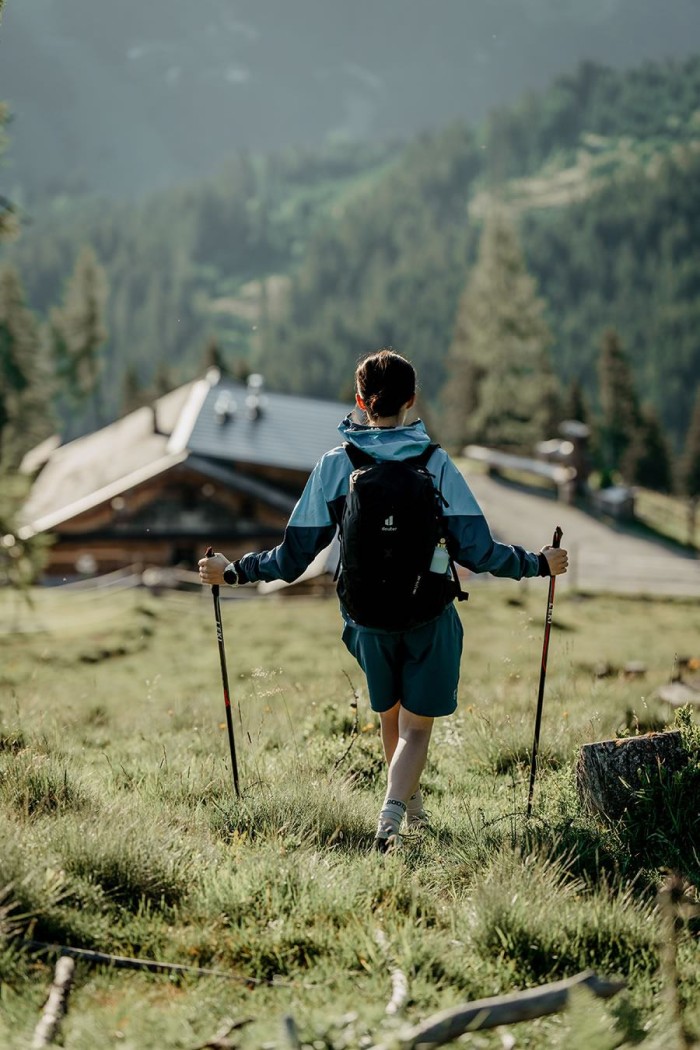 Person wandert auf einer Wiese mit Wanderstöcken und einem Rucksack in Richtung Hütte