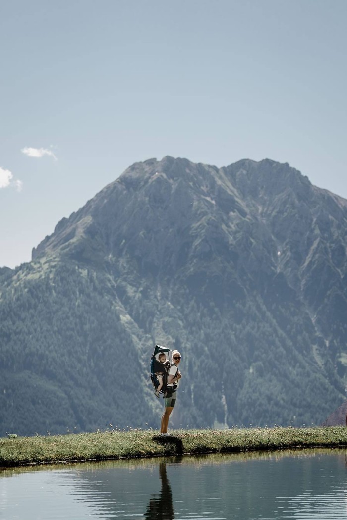 Person trägt Kind in Trage, vor großer Bergkulisse und neben einem Wasserbecken stehend