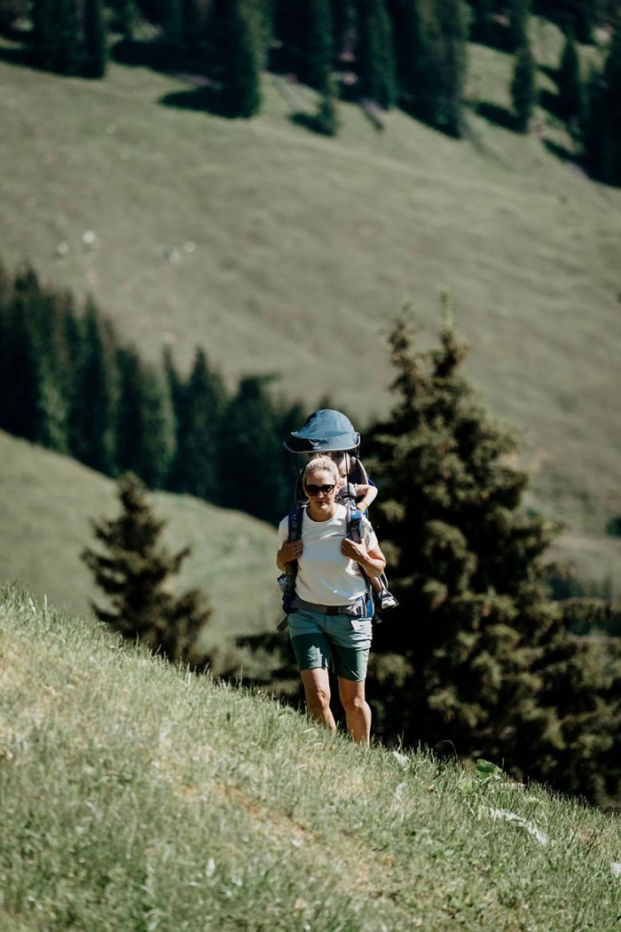 Person wandert mit einem Kind in einer Trage in einer bergigen Grünlandschaft
