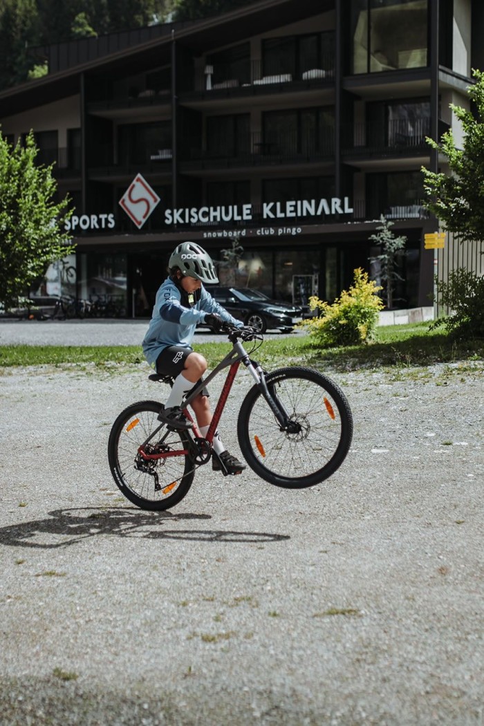 Ein Kind macht einen Wheelie auf einem Fahrrad vor einer Skischule