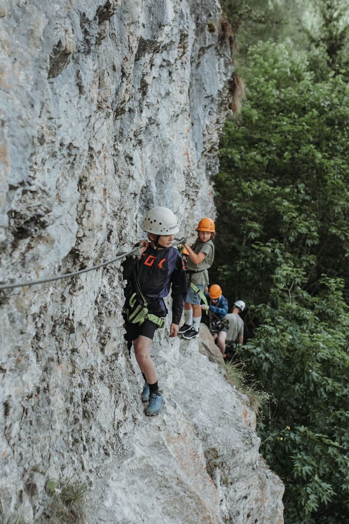 Kinder klettern gesichert an einem Felsvorsprung entlang, umgeben von grüner Vegetation