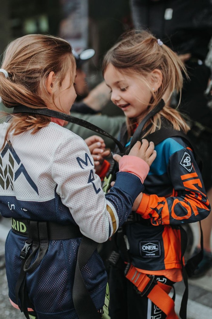 Zwei Kinder in bunten Jacken helfen sich gegenseitig beim Anlegen von Klettergurten