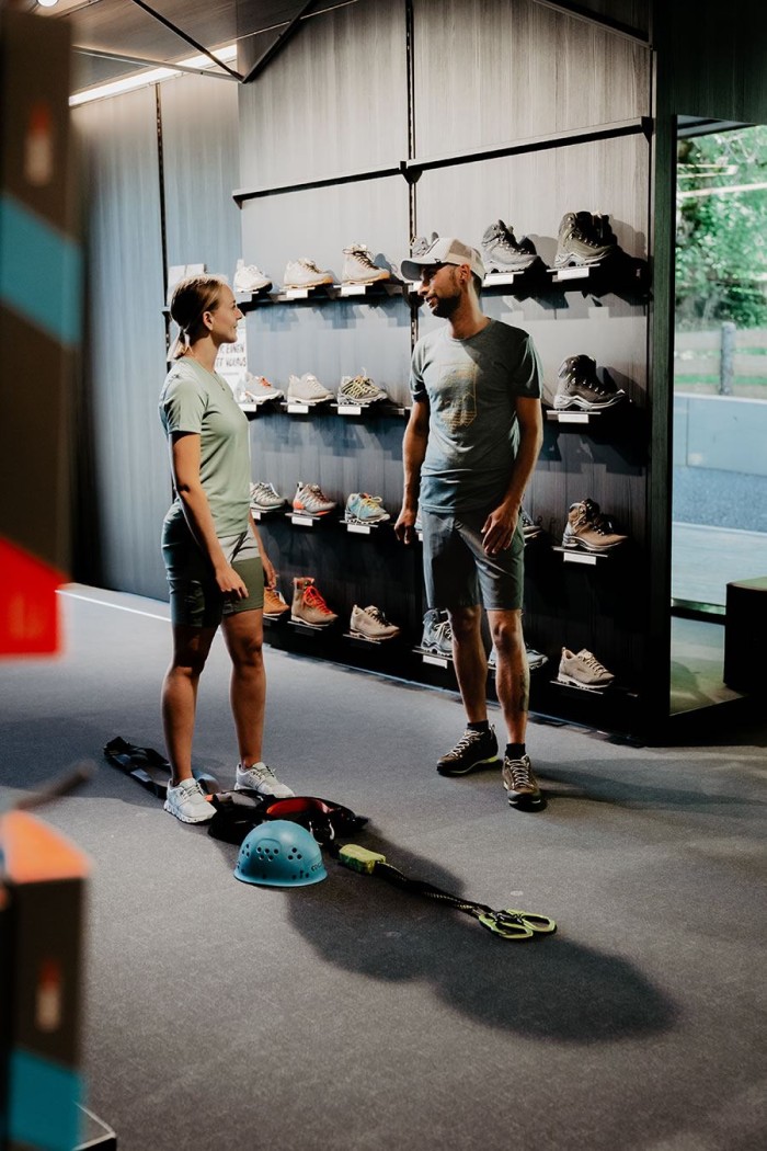 Zwei Personen stehen in einem Geschäft vor einem Regal voller Schuhe, mit Kletterausrüstung am Boden