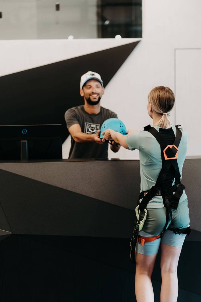 Person in Kletterausrüstung erhält Helm von Mitarbeiter am Empfangstresen