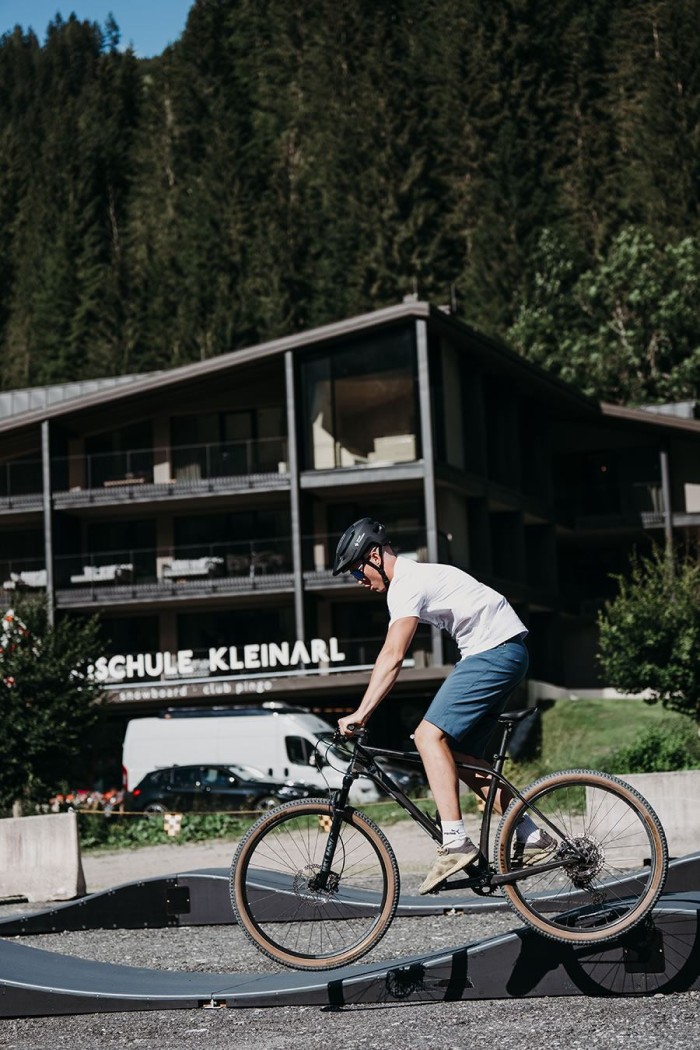 Person auf Fahrrad fährt über wellige Strecke vor Gebäude in bergiger Landschaft