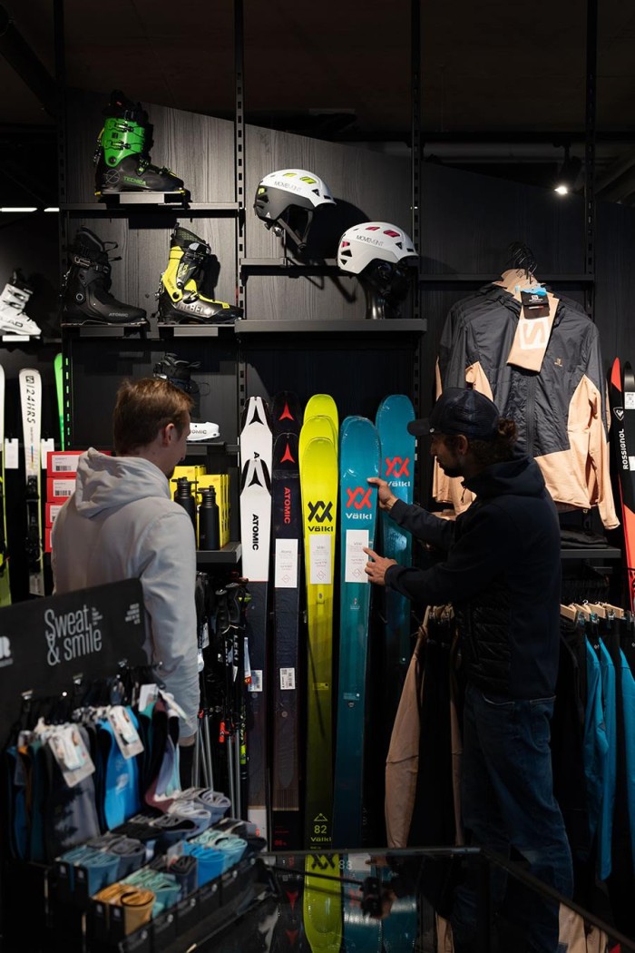 Zwei Männer betrachten Skier in einem Sportgeschäft mit Zubehör wie Helmen und Jacken